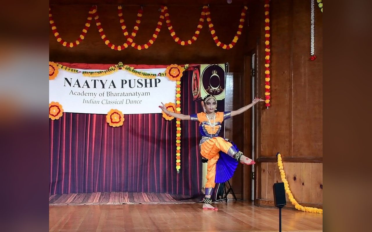 A dancer in an orange and blue costume leaps across the stage beneath the Naatya Pushp banner and marigold garlands