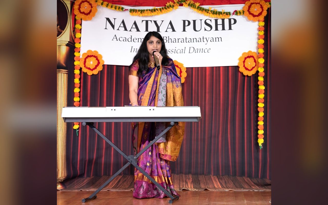 A vocalist in a purple sari sings at a keyboard in front of the Naatya Pushp banner