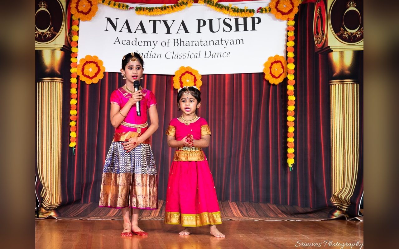 Two young dancers in pink costumes speak to the audience beneath the Naatya Pushp banner