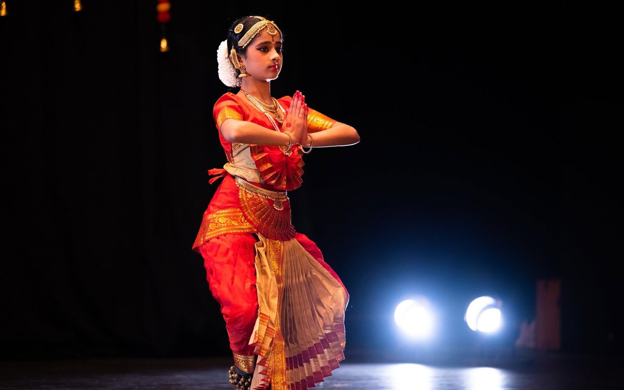A dancer in a red and gold costume performs in a spotlight on a dark stage