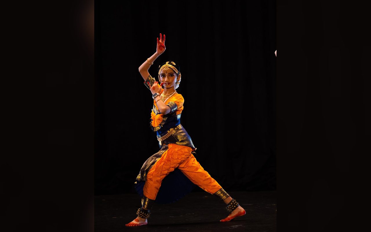 A dancer in orange and blue lunges with one arm raised high