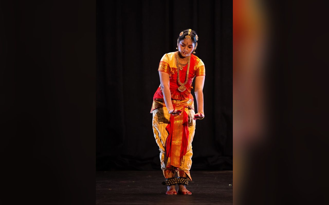 A dancer in orange and red stands poised in an opening position