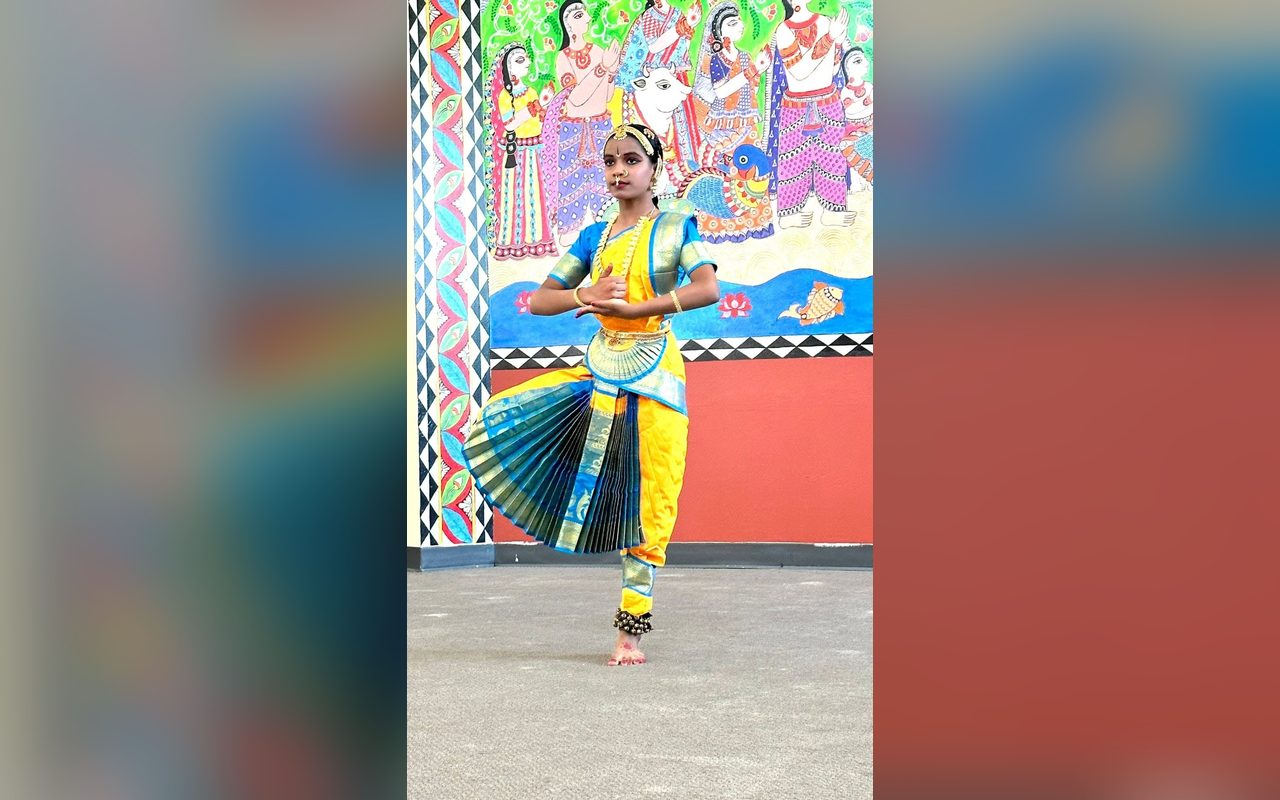 A young dancer in a yellow and blue costume performs before a colorful mural of Indian folk art