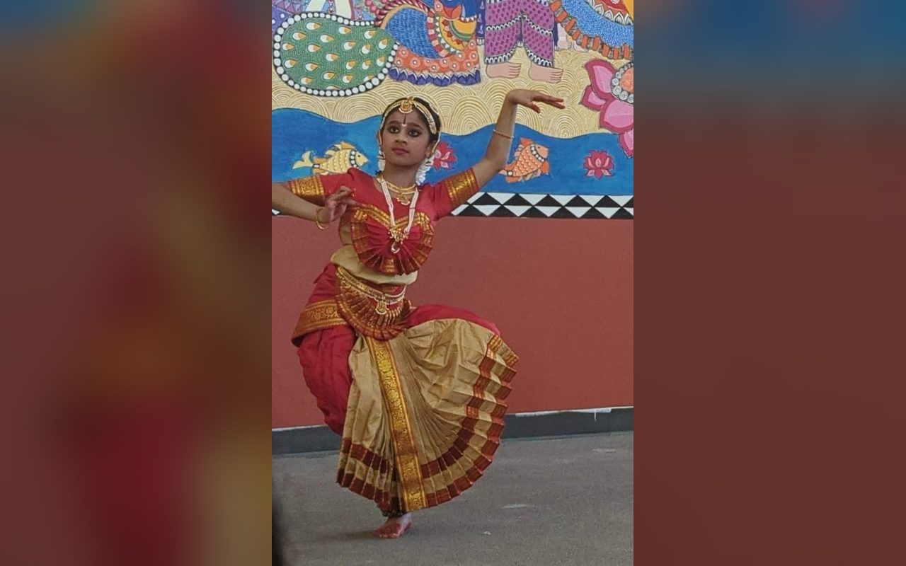 A dancer in a red and gold costume holds a pose with one arm extended before a painted mural