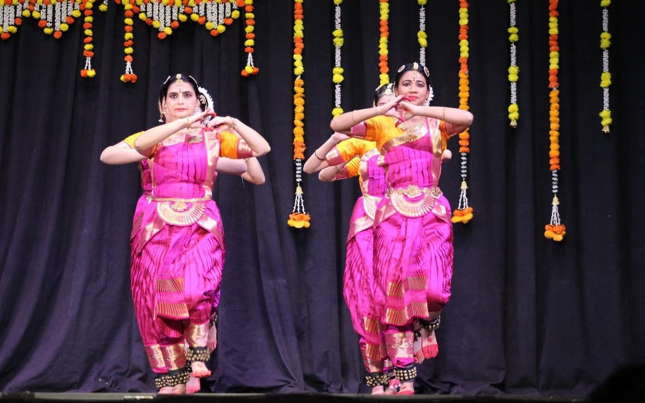 Dancers in pink and gold costumes perform in pairs before a curtain of marigold garlands