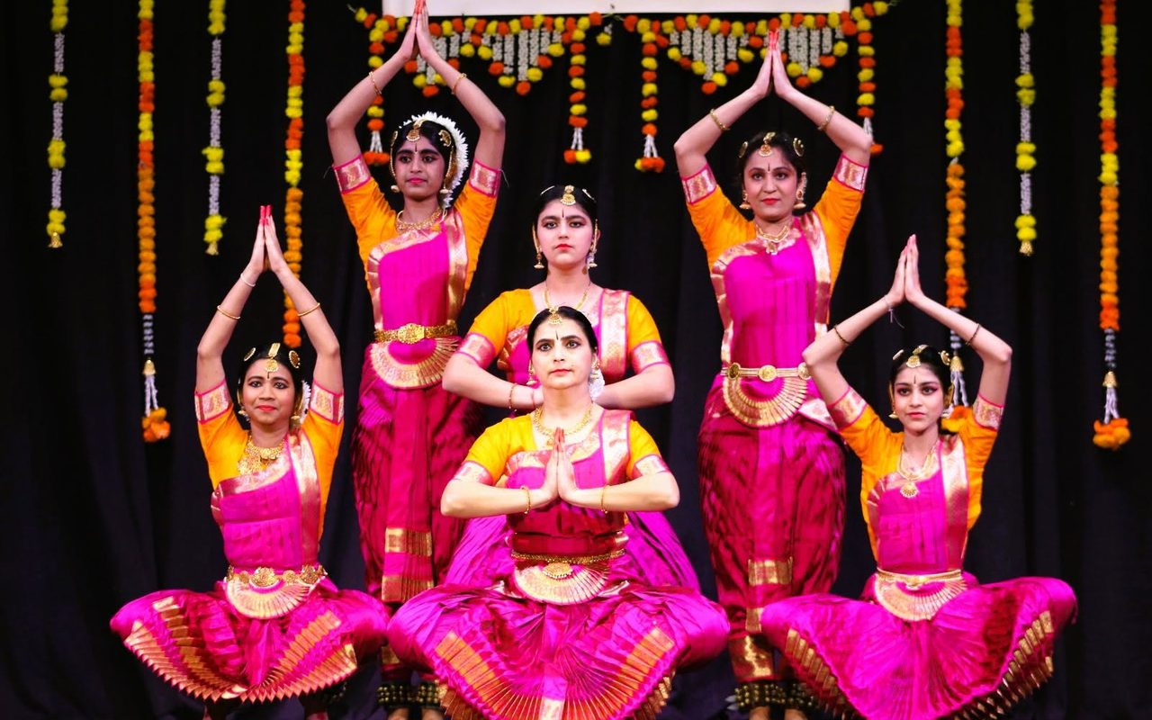 A group of dancers in pink costumes forms a tiered pose with palms joined overhead