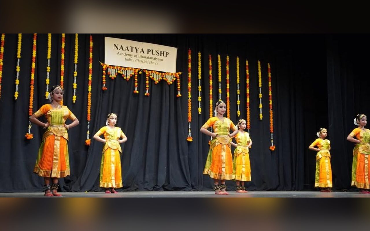 Young dancers in yellow and orange costumes perform in a line beneath the Naatya Pushp banner