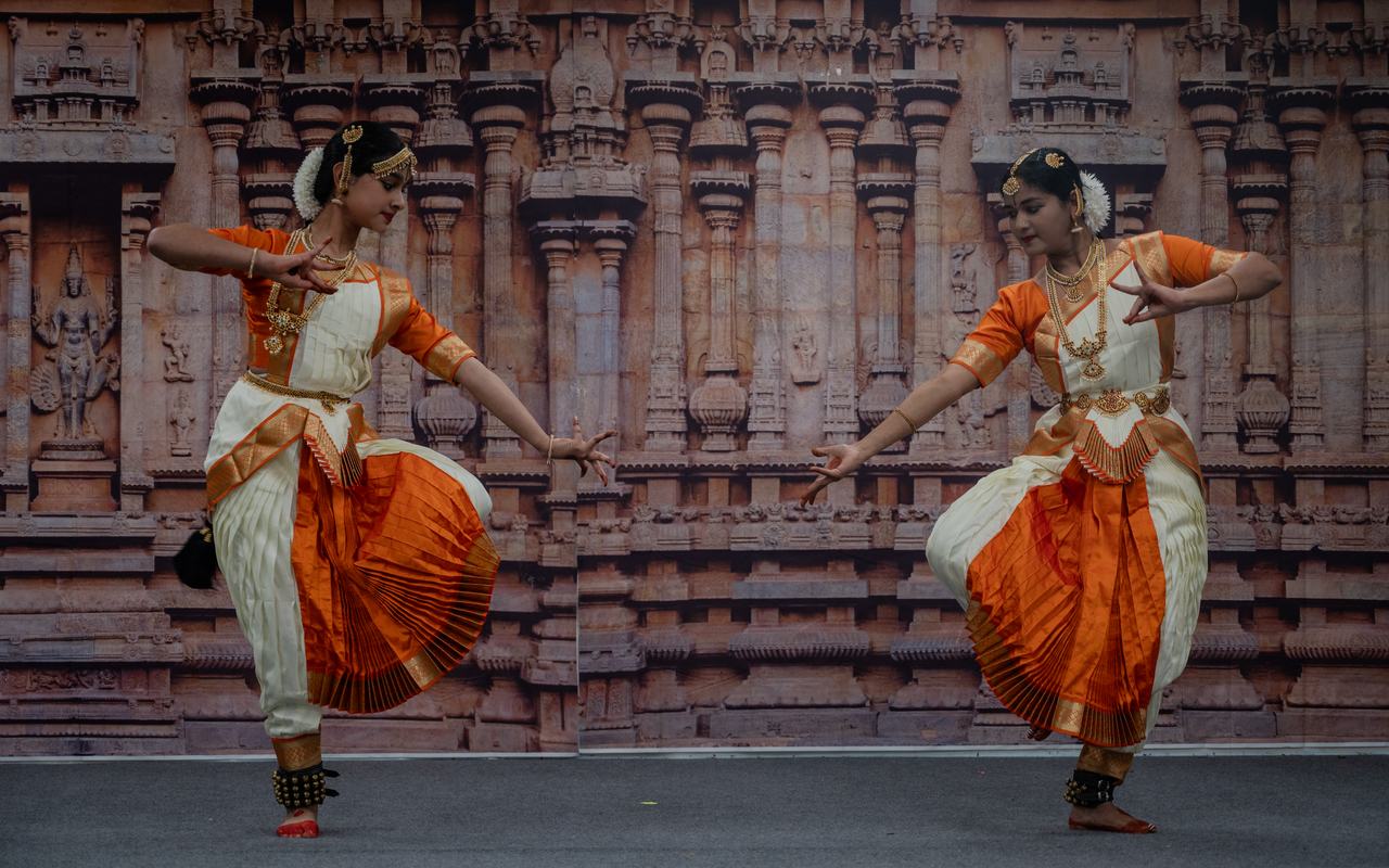 Two dancers in orange and white costumes perform facing each other before a temple-carving backdrop