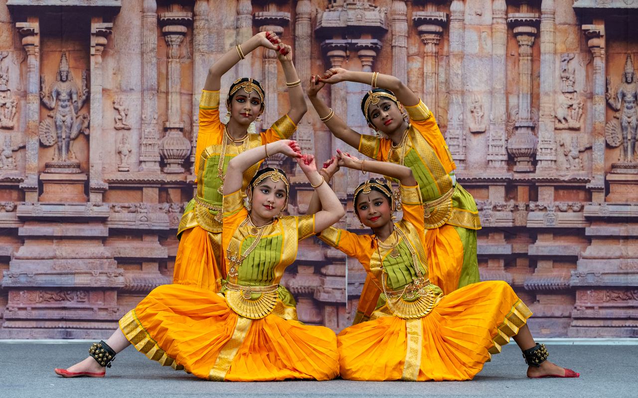 Four dancers in yellow costumes hold a symmetrical pose, arms framing their faces