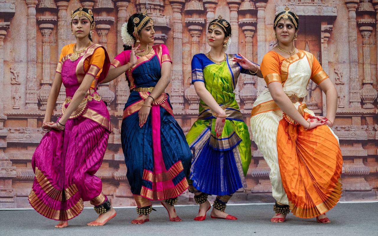 Four dancers in pink, blue, green and orange costumes pose together before a temple-carving backdrop