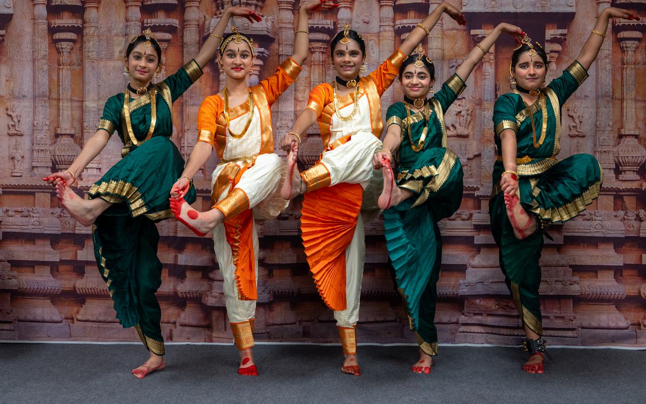 Five dancers in green and orange costumes balance on one leg in unison