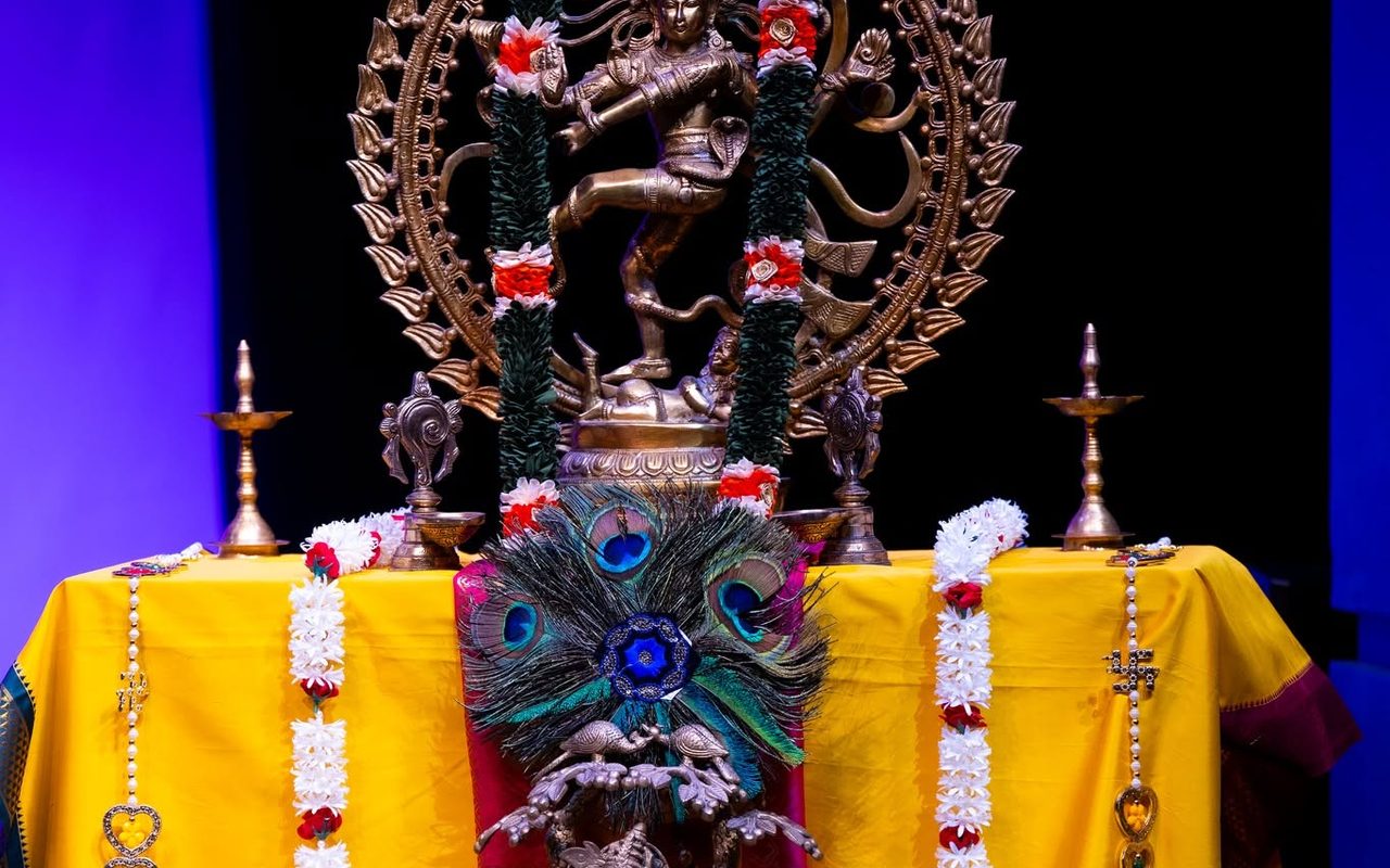 The stage altar: a Nataraja statue garlanded with flowers, brass lamps, and a peacock-feather Krishna idol on yellow silk