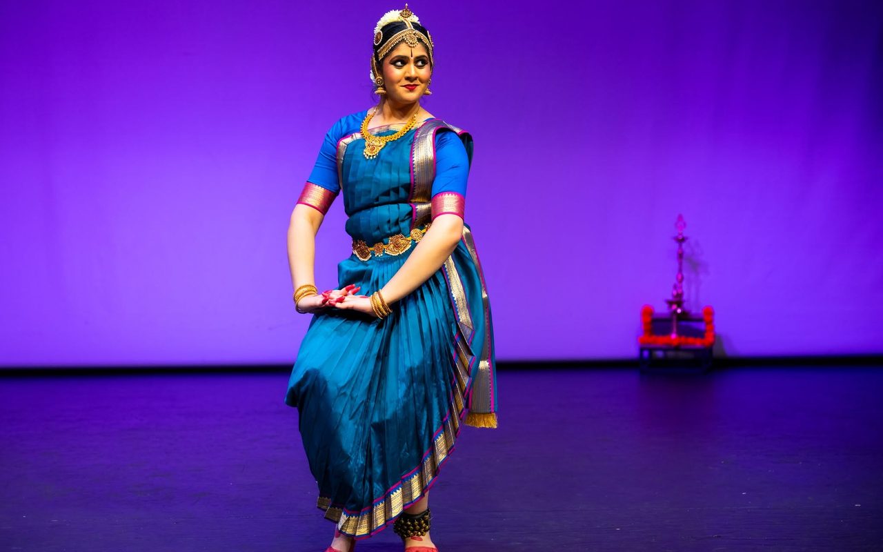 Shreya, in a teal and gold Bharatanatyam costume, holds a poised stance on a violet-lit stage