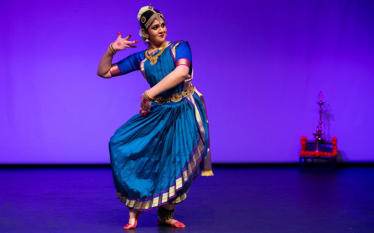 Shreya, in a teal costume, strikes a graceful pose with one hand raised, a brass lamp glowing behind her