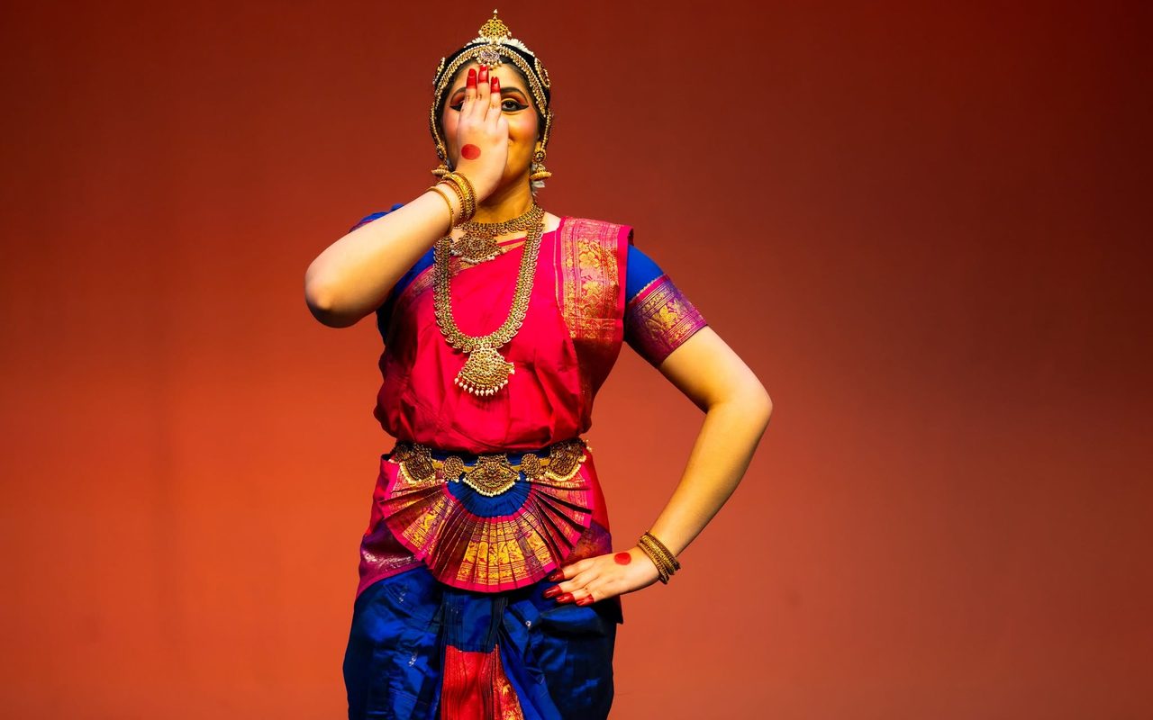 Shreya, in a pink and blue costume, performs an expressive gesture against a deep red backdrop