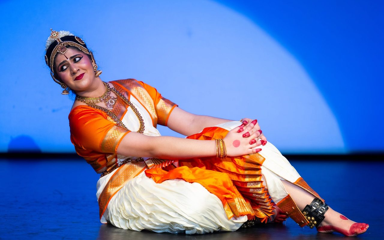 Shreya, in an orange and cream costume, reclines on the stage with a playful sideways glance