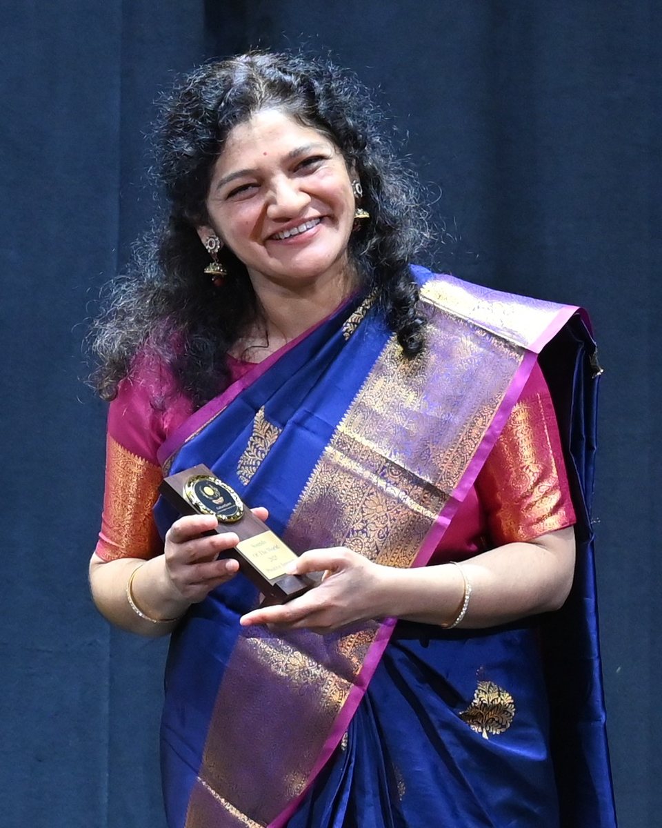 Guru Smt. Olympia Mannur, smiling, in a blue and gold silk sari, holding an award on stage